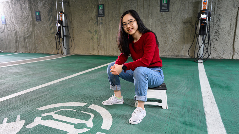 Elaine Siyu Liu sits on a stool inside a parking garage. The ground is painted green and there is an electric car logo. Plus, in the background are electric are charging stations.